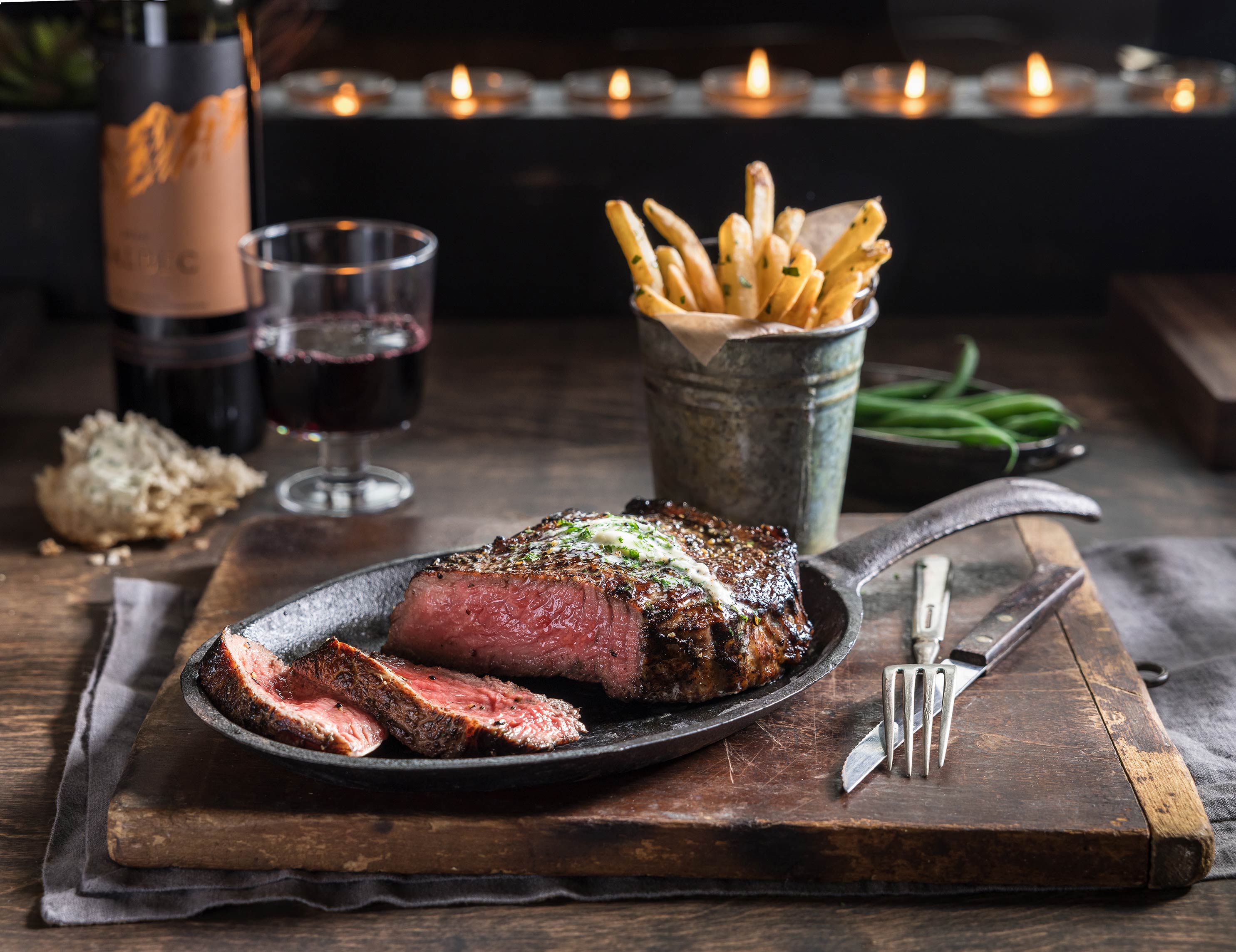 Glanger Photography | Medium rare steak on a skillet with herb butter, fries and green beans.  Rustic restaurant with wine, torn bread and candles in the background. 