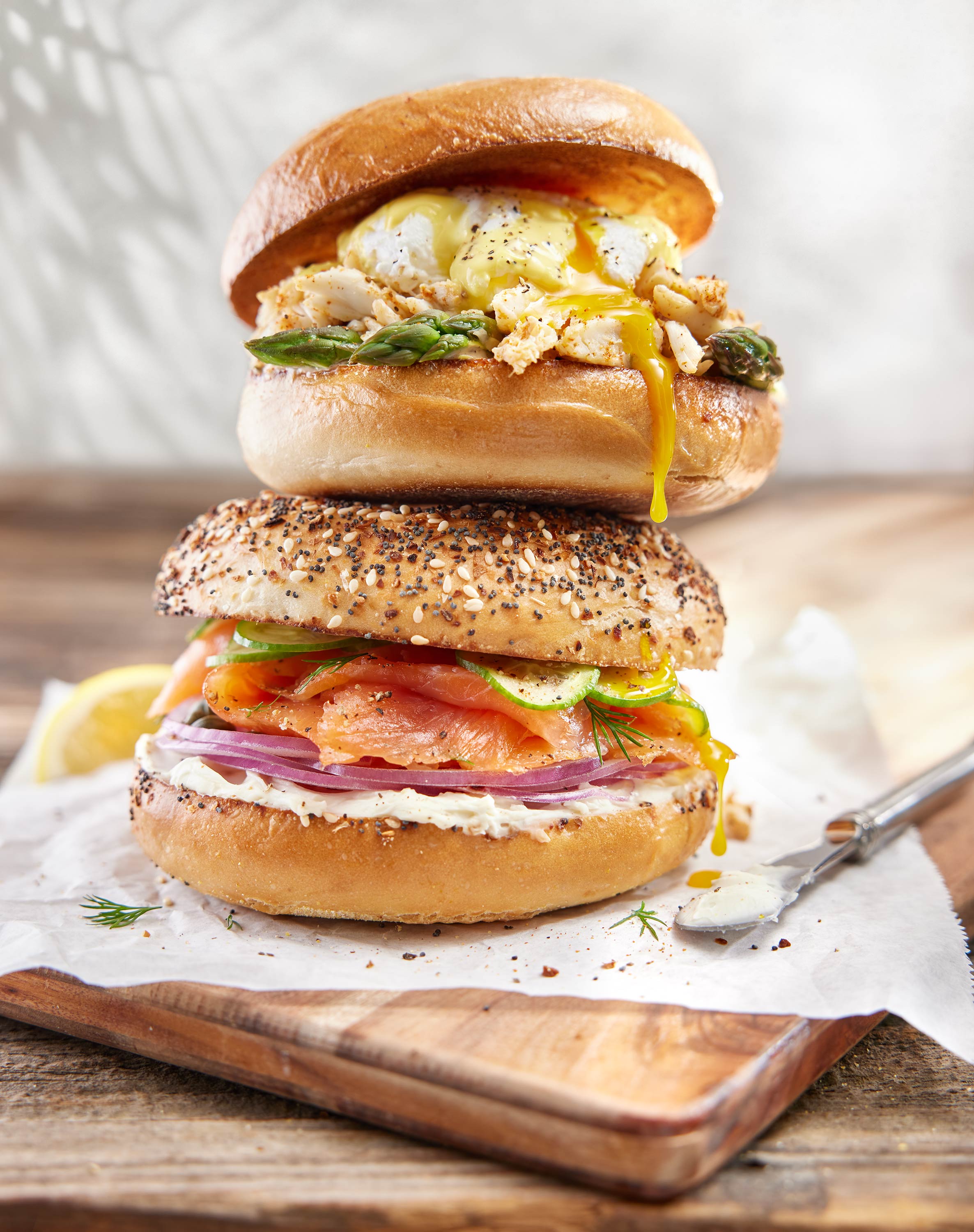 Double stack of bagels. Bottom bagel with lox, cream cheese, thinly sliced cucumber, red onions and black pepper.  Top Bagel with a hollandaise egg with crab and asparagus.  Yolk  is dripping down. 