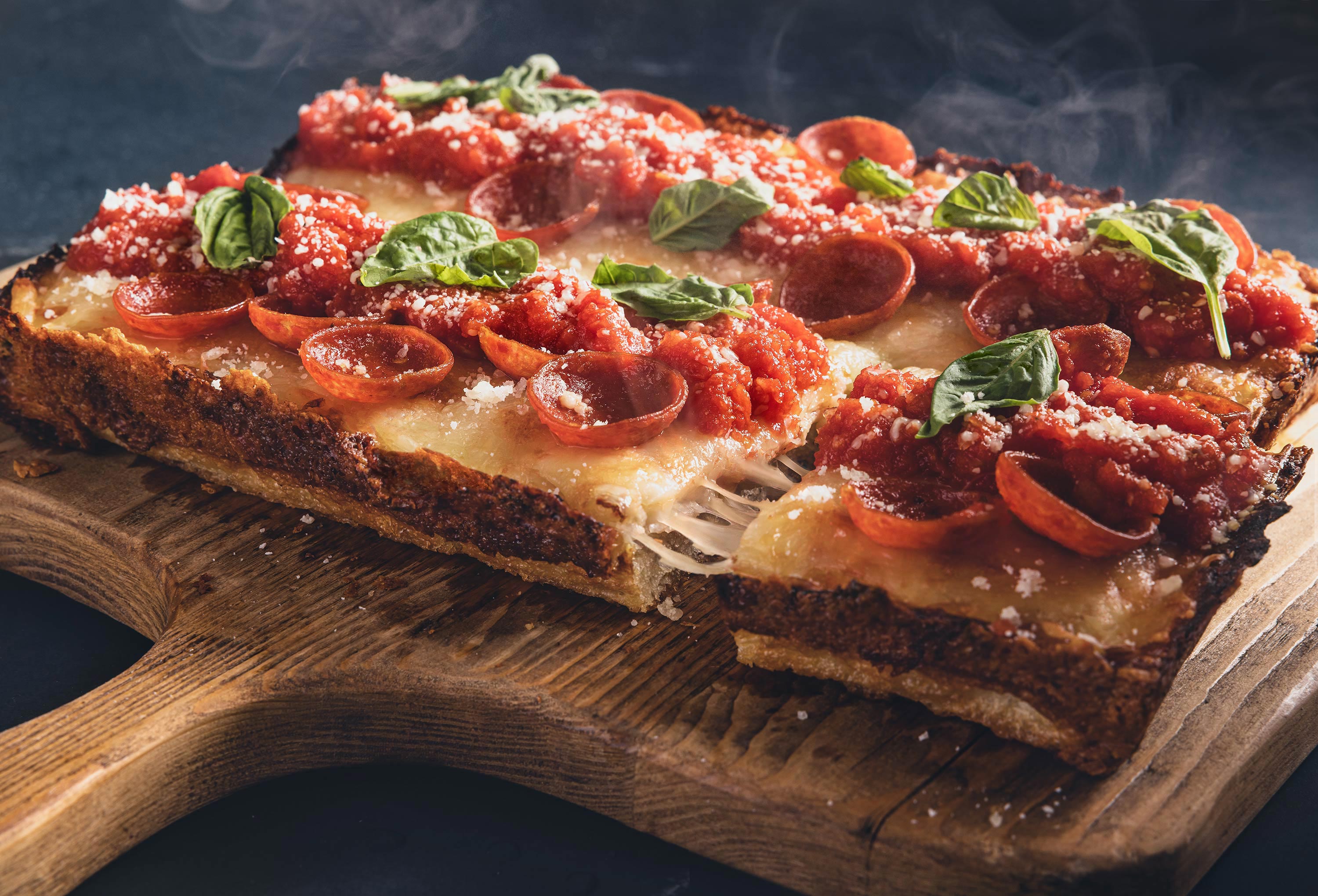 Deep dish rectangular pepperoni pizza with basil and one slice being taken out showing a cheese pull.  Pizza sits on a rustic paddle style cutting board